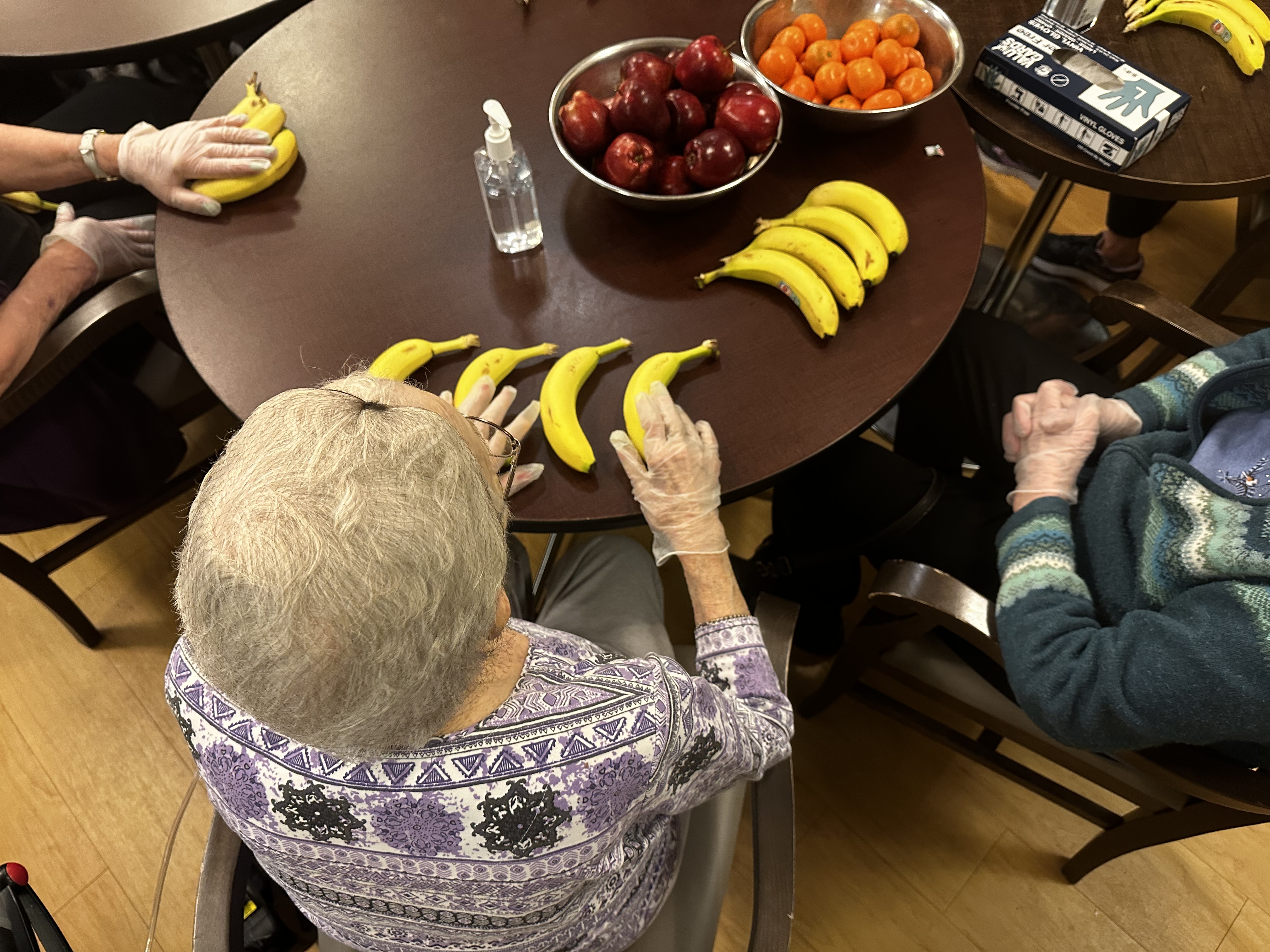 An aerial image looks down on three seniors arranging fruit on a brown round table.